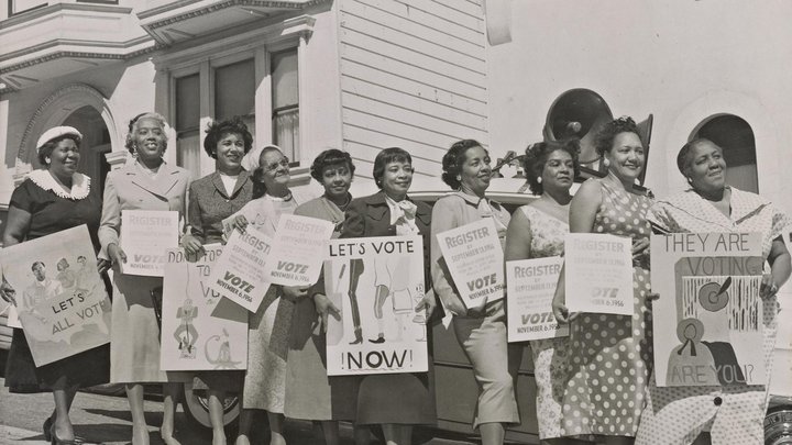 A black and white photo from 1956. African American women lined up in "their Sunday Best" holding signs saying, "Let's Vote Now."