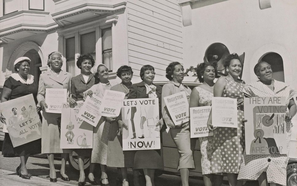 A black and white photo from 1956. African American women lined up in "their Sunday Best" holding signs saying, "Let's Vote Now."