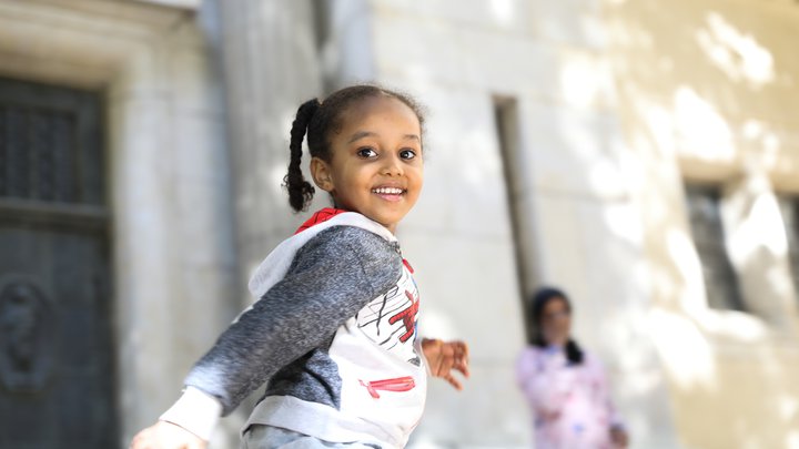 Young girl with her hair in braids and wearing a red, white and grey sweatshirt smiles at the camera, while running, with her mother in the background wearing a pink dress and dark headscarf, talking on the phone.