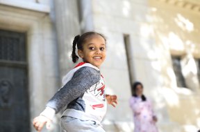 Young girl with her hair in braids and wearing a red, white and grey sweatshirt smiles at the camera, while running, with her mother in the background wearing a pink dress and dark headscarf, talking on the phone.