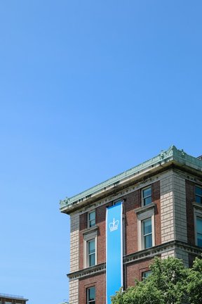 Columbia University building in the lower right-hand corner with a Columbia Blue banner. Entire image is shared on a blue sky background.