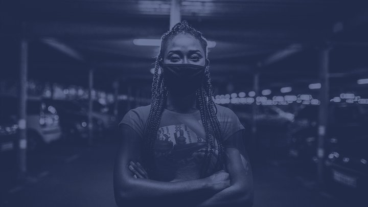 A women with braids, wearing a surgical face mask, stands in a garage full of cars with her arms crossed.