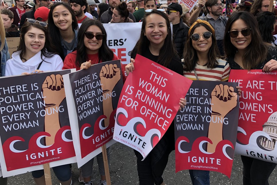 Six young women stand in a crowd, holding poster and signs from IGNITE, promoting women's political power. The posters are red and grey, with an image of a wave at the bottom. Their t-shirts are different colors and four of them are wearing sunglasses.