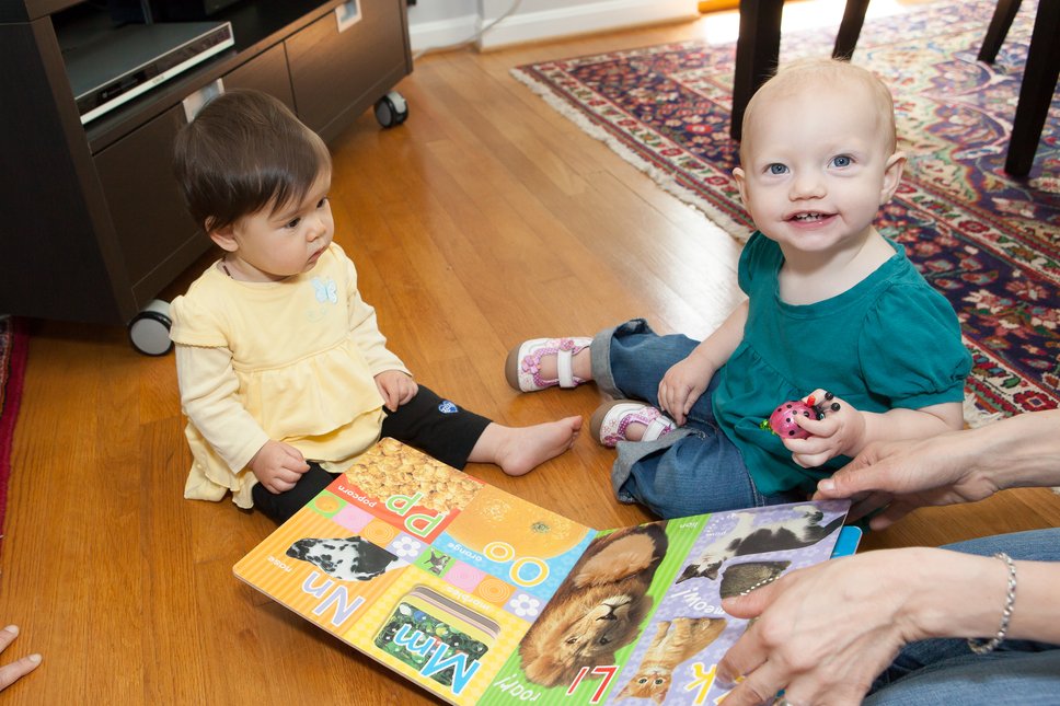 Two children sit on a hardwood floor playing with a book of the alphabet and a toy ladybug