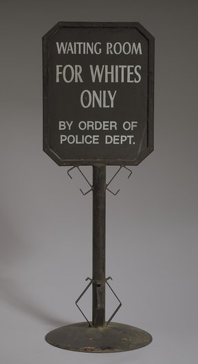 A metal double-sided black railroad sign that reads: [WAITING ROOM / FOR WHITES / ONLY / BY ORDER OF / POLICE DEPT.] in white paint. The writing is on a framed square plaque at the top of the sign. The sign post is round and has metal work at the top wher