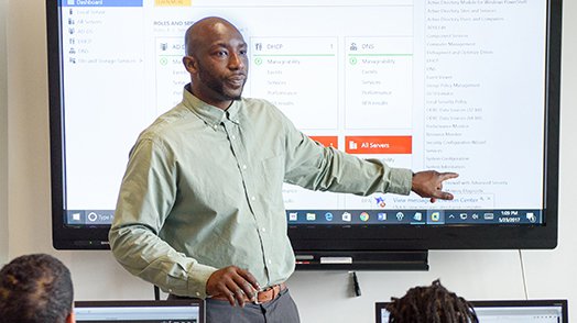 An African-American man stands at the front of a room, teaching a technology class. Dressed in a green shirt and grey slacks, he is pointing at a screen.