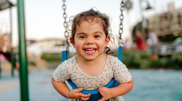 Celebrating the Build a World of Play Challenge with this image of a young girl with curly wet hair smiling at the camera. She is leaning on a blue swing and wearing a yellow dress with blue dots. In the blurry background is a playground.