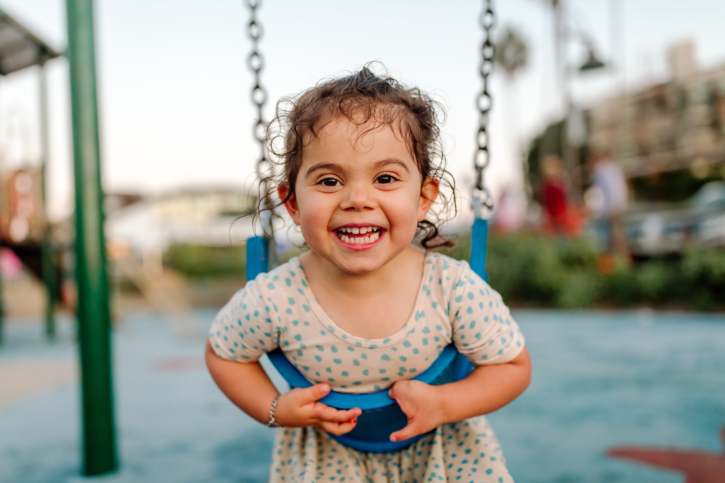 Celebrating the Build a World of Play Challenge with this image of a young girl with curly wet hair smiling at the camera. She is leaning on a blue swing and wearing a yellow dress with blue dots. In the blurry background is a playground.